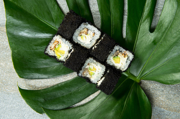 Bovenaanzicht van Japanse sushi roll met unagi paling, roomkaas, avocado verpakt in zwarte kaviaar (vliegende viskuit Tobiko) geserveerd op exotische monstera blad op grijze stenen achtergrond. voedselkunstconcept - Foto, afbeelding