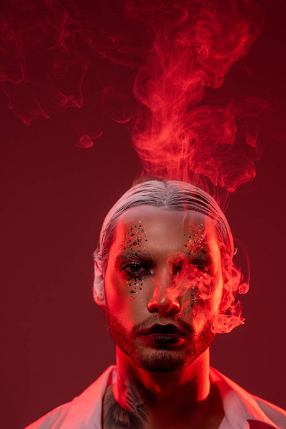 Retrato de un joven carismático serio con maquillaje brillante y barba contra un fondo rojo aislado, humo alrededor de su cara y por encima de la cabeza - Foto, Imagen