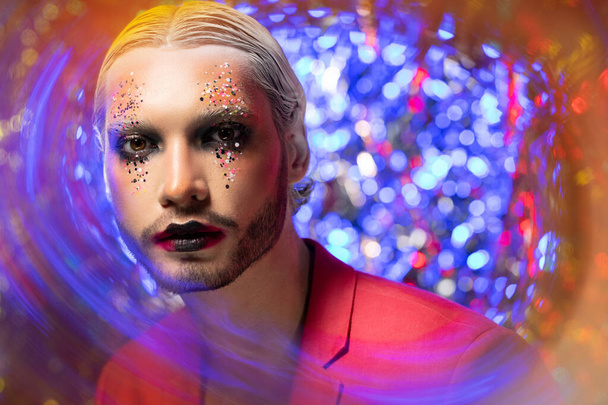 Portrait of serious young bearded man with New Year womanish makeup posing against glitter background - Foto, afbeelding