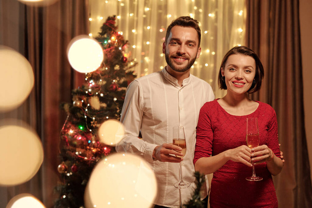 Mosolygó fiatal pár portréja ünnepi öltözetben pezsgőfuvolával díszített fán, karácsonyi fények hatása - Fotó, kép