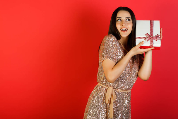Hübsche junge brünette Frau in beigem Kleid schüttelt Geschenkbox, um herauszufinden, was drin ist. Roter Hintergrund mit Leerraum für Text. - Foto, Bild