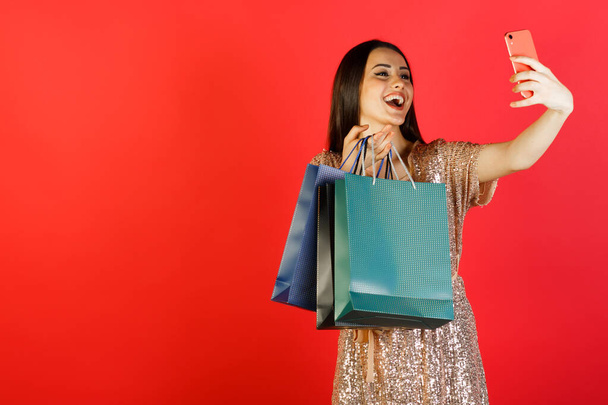 Portrait of smiling woman in dress holding packages bags with purchases after shopping and doing selfie isolated on red pastel background. Copy space for advertisement. - Photo, Image