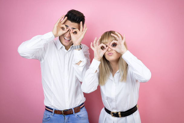 Молода пара дівчини і хлопець над ізольованим рожевим тлом робить ОК жест шокований усміхненим обличчям, око дивиться через пальці
 - Фото, зображення