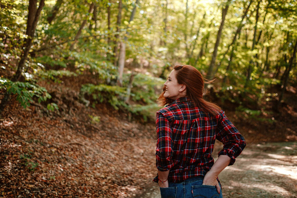 Ταξιδιώτρια hipster γυναίκα στέκεται μόνη της στο δάσος φθινόπωρο σε καρό πουκάμισο και μπλε τζιν. Κρύο καιρό, φθινοπωρινά χρώματα. Έννοια της περιπλάνησης. - Φωτογραφία, εικόνα