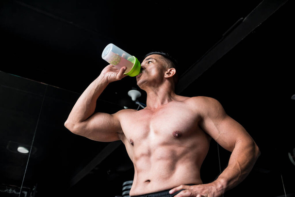 A fit asian man drinks BCAA or sports drink as a pre workout drink before an intense workout. Gym or fitness club setting. - Фото, изображение
