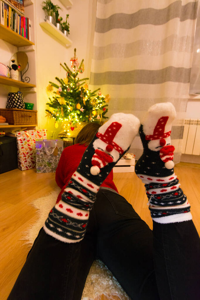 A vertical shot of a lady wearing Christmas socks sitting near the Christmas tree and presents - Photo, image