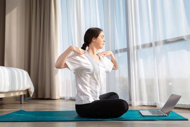 Eine junge Frau in Sportbekleidung sitzt und macht Yoga auf Online-Unterricht. Indoor. Seitenansicht. Das Konzept des Online-Trainings zu Hause. - Foto, Bild