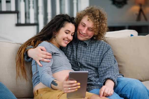 Good-looking pregnant couple relaxing on the comfortable couch at home and watching tv shows on tablet pc. - Foto, Bild
