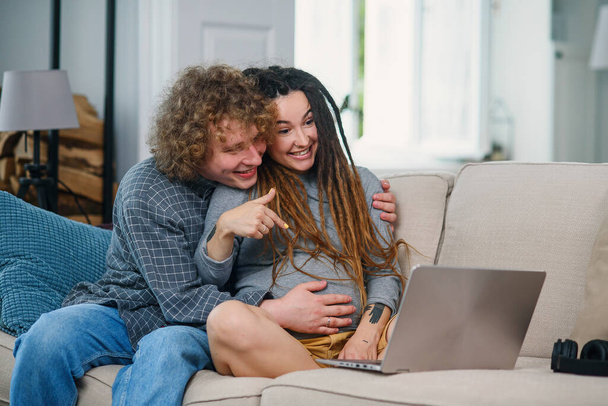 Loving man sits with his pregnant wife with stylish dreadlocks on the couch and having video chat on computer. - Fotó, kép