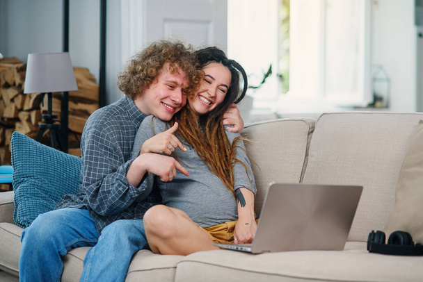 Loving man sits with his pregnant wife with stylish dreadlocks on the couch and having video chat on computer. - 写真・画像