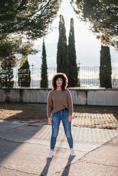 A beautiful female with curly hair in a park wearing a beige sweater and jeans with white sneakers - Photo, image