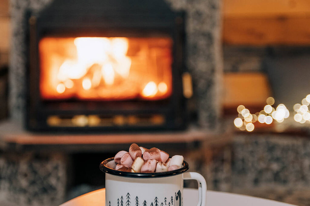 A mug of cocoa or hot chocolate with powdered marshmallows placed on a table by the fireplace - Foto, immagini