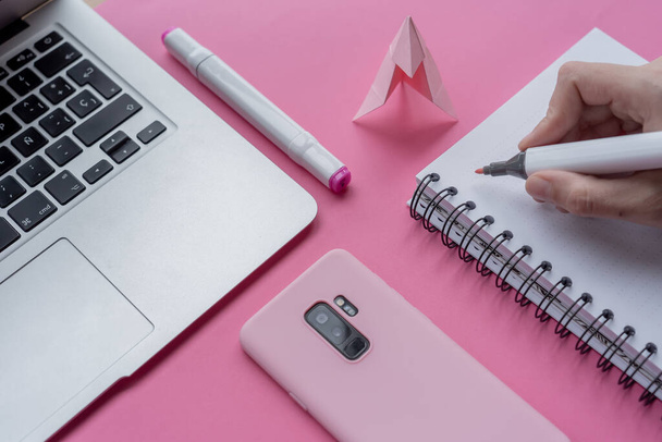 A hand holding marker with a notebook, laptop, phone, and swan origami on pink background - Foto, Imagen