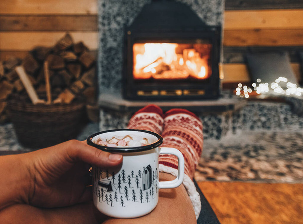 A woman holding a mug of cocoa or hot chocolate with powdered marshmallows by a warm fireplace in winter - Photo, image