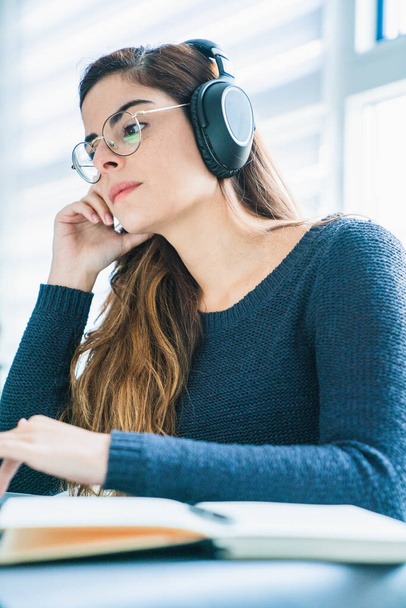 Μια νεαρή Ισπανίδα με γυαλιά που ακούει μουσική με ακουστικά και δουλεύει στον υπολογιστή. - Φωτογραφία, εικόνα