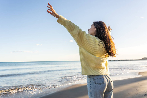 A shallow focus of a female wearing a yellow sweater and standing on a beach with open arms in Benicassim, Spai - 写真・画像