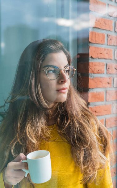 A young Hispanic female in a yellow shirt drinking coffee and looking through the window - Foto, imagen