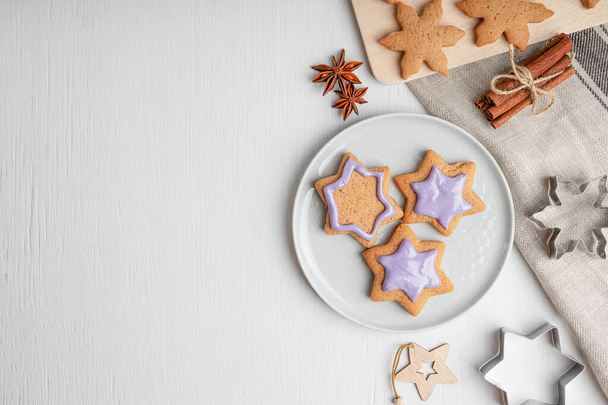 Portion of sweet savory gingerbread cookies with blue glaze served on gray plate on white wooden background with pastry cutters, textile towel and cinnamon. Image with copy space, flat lay - Fotó, kép