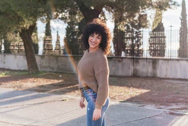 A beautiful female with curly hair in a park wearing a beige sweater and smiling - Fotó, kép