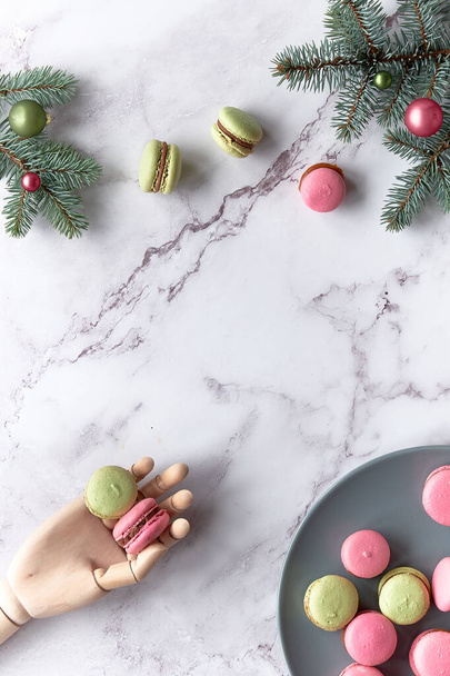 Pink and mint green macaroons in wooden hand and on grey plate. Tasty creative dessert in green and red on marble table with fir twigs. Happy winter holidays - Φωτογραφία, εικόνα