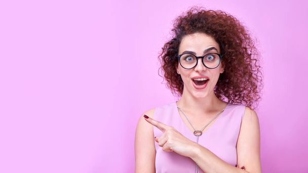 Portrait of amazed caucasian curly girl wearing glasses points finger to the side on copy space isolated in pink studio background - 写真・画像