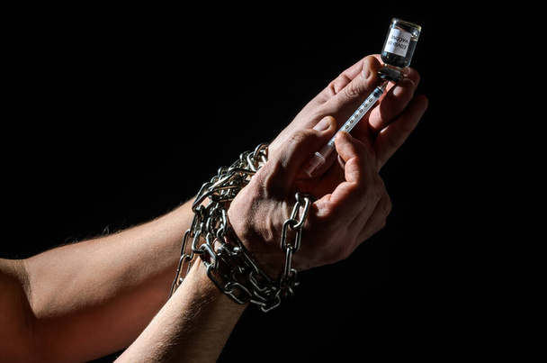 Human hands chained in chains are taking an injection into a syringe from an ampoule with a covid-19 vaccine isolated on a black background - Фото, зображення