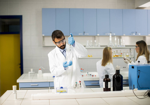 Όμορφος νέος ερευνητής με προστατευτικά ενδύματα εργασίας που στέκεται στο εργαστήριο και αναλύει τη φιάλη με υγρό - Φωτογραφία, εικόνα
