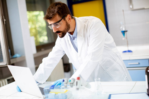 Fešák mladý vědec v bílém laboratorním plášti pracuje pomocí notebooku, zatímco sedí v laboratoři - Fotografie, Obrázek