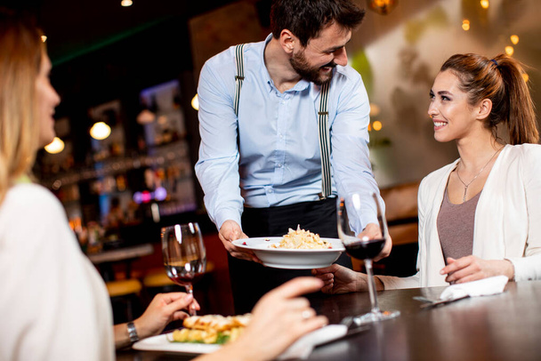 Komea nuori tarjoilija tarjoilee ruokaa naispuolisille asiakkaille ravintolassa - Valokuva, kuva