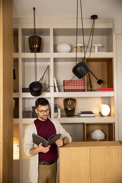 Handsome young man in casual clothes and with eyeglasses reading a book at home - Fotó, kép