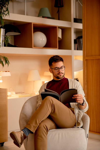 Handsome young man in casual clothes and with eyeglasses reading a book at home - Foto, Imagem