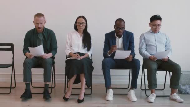 Full shot of four nervous diverse job applicants sitting on chairs in hallway, waiting for their turn, unrecognizable manager inviting young African man - Footage, Video