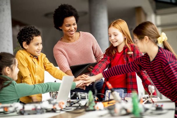 Grupa szczęśliwych dzieci ze swoją afroamerykańską nauczycielką fizyki z laptopem programującą zabawki elektryczne i roboty w klasie robotyki - Zdjęcie, obraz