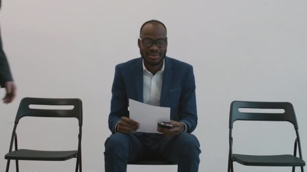 Mittlere Langzeitaufnahme eines afrikanischen Geschäftsmannes, der auf einem Stuhl im Flur sitzt, Papier in den Händen hält, unkenntliche Menschen, die an ihm vorbeigehen, asiatische Kollegen, die ihn grüßen - Filmmaterial, Video