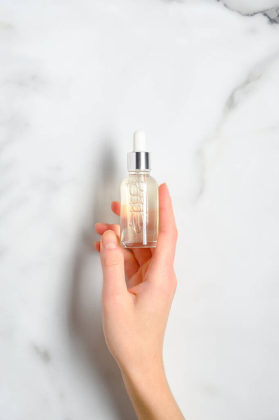 Transparent dropper bottle with serum lotion or essential oil in female hand over marble table. Skin care cosmetics concept. - Photo, image