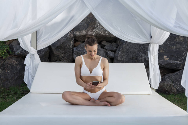 Mujer alegre con bikini blanco sentado con las piernas cruzadas en la cama con dosel y navegar por Internet en el teléfono móvil mientras se relaja durante las vacaciones de verano - Foto, Imagen