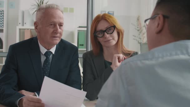 Chest-up over shoulder of elderly Caucasian male manager and red-haired businesswoman sitting together, talking, looking interestedly at unrecognizable candidate on job interview - Séquence, vidéo