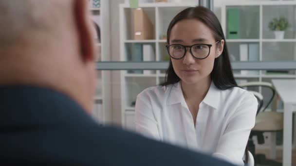 Borst-up over schouder van slimme Aziatische vrouwelijke kandidaat in wit shirt en bril zitten in het kantoor, praten met onherkenbare manager op sollicitatiegesprek - Video