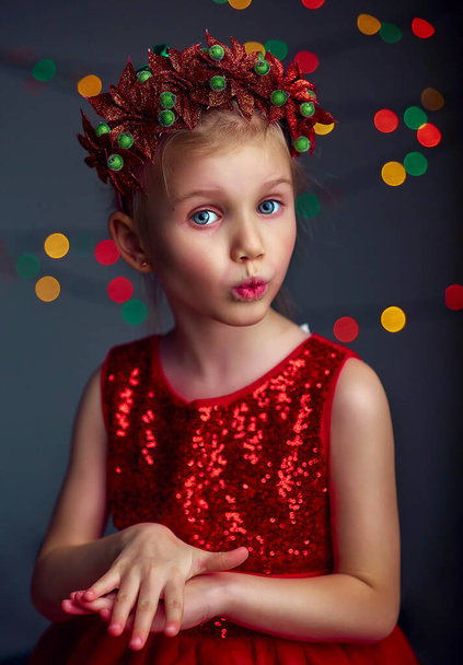 Karácsonyi portré boldog fiatal lány visel piros flitteres ruhát és karácsonyi koszorú. Szőke haj és kék szem pózol és bolondozik a stúdióban szürke háttér megvilágítás és boke fények. - Fotó, kép