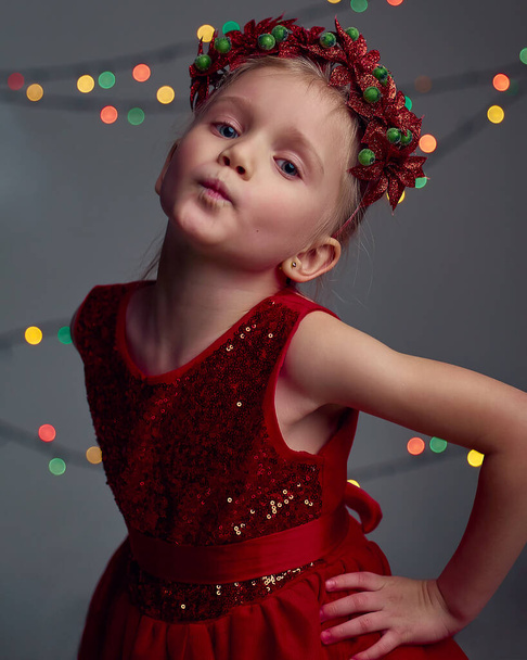 Різдвяний портрет щасливої молодої дівчини в червоній сукні з шпинатом та різдвяному вінку. Блондинка волосся і блакитні очі позує і дурень навколо в студії на сірому фоні з підсвічуванням і вогнями
. - Фото, зображення