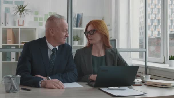 Chest-up of aged Caucasian businessman and ginger-haired middle-aged female manager sitting together at desk in office talking, unrecognizable job candidate joining them - Кадры, видео
