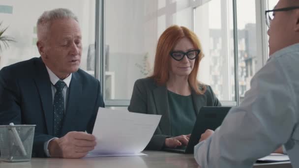 Medium close-up of senior Caucasian businessman and red-haired middle-aged HR manager sitting in office, interviewing unrecognizable candidate. Man reading from paper, talking, female colleague typing - Záběry, video
