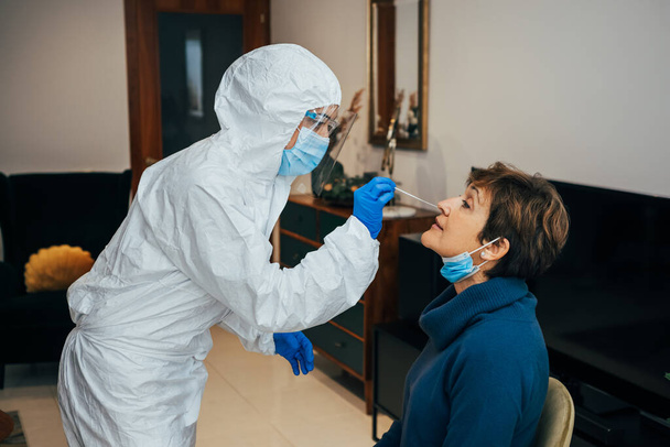 Gesundheitsexpertin mit PSA, Gesichtsmaske und Schutzschild, die einer älteren Patientin bei einem Hausbesuch einen Nasenabstrich vorführt. Antigen-Schnelltest zur Analyse von Nasenproben auf Covid-19, Coronavirus-Pandemie. - Foto, Bild