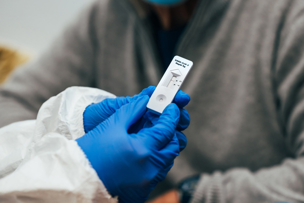 Nahaufnahme von Arzthänden mit Schutzhandschuhen und PSA-Anzug, die einem älteren Patienten ein Testgerät zeigen. Überprüfung eines Antigen-Schnelltests für SARS Covid-19. - Foto, Bild