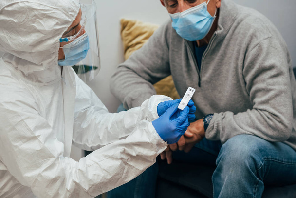 Mediziner im PSA-Anzug, Maske, Gesichtsschutz, der dem Patienten das negative Ergebnis für Covid-19 zeigt. Antigen-Test bei einem älteren Patienten während eines Hausbesuchs. Coronavirus-Pandemie. - Foto, Bild