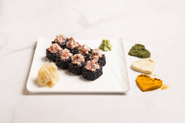 Zijaanzicht van Japanse zwarte rijst sushi roll met unagi paling geserveerd met gember, wasabi op vierkante plaat. Drie Hart silhouetten van basilicum, knoflook, kurkuma poeder specerijen op witte marmeren achtergrond - Foto, afbeelding
