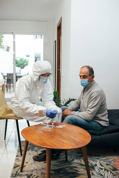Professioneller Arzt im Anzug, Gesichtsmaske, Gesichtsschutz, der den Antigen-Test vorbereitet. Ein älterer erwachsener Patient wartet zu Hause auf die Entnahme von Nasenkulturproben während der Coronavirus-Pandemie. - Foto, Bild