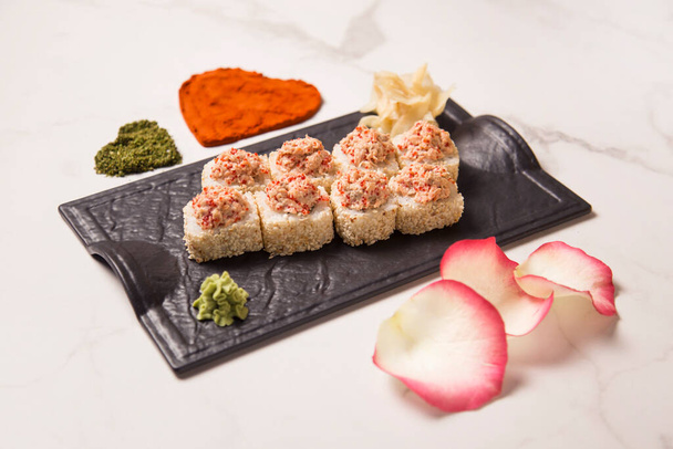 Boční pohled na japonský sushi rohlík s pečeným lososem a tobiko létající rybí jikry podávané na černém talíři s wasabi a zázvorem. srdce tvary kurkuma a bazalky koření a okvětní lístky na mramorovém pozadí - Fotografie, Obrázek