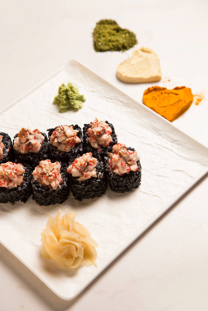 Primer plano del rollo de sushi de arroz negro japonés con anguila unagi servida con jengibre, wasabi en plato cuadrado. Tres siluetas de corazón de albahaca, ajo, cúrcuma en polvo especias sobre fondo de mármol blanco - Foto, imagen