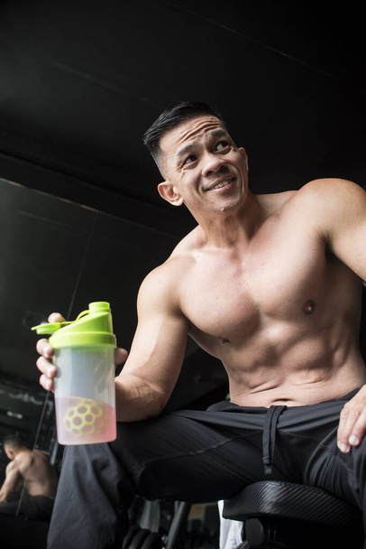 A fit and handsome middle aged male sitting on a bench and holding a sports drink after a vigorous workout at the gym. In his early 40s but very youthful looking. - Photo, Image